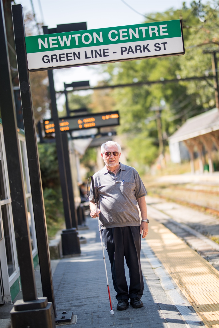 An older adult holding his white mobility cane waits for a train at the Newton Centre train station.