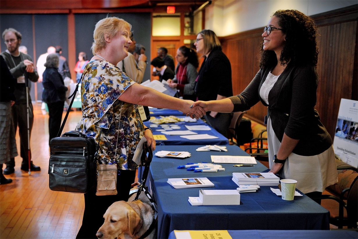 A job seeker reaches across the table and shakes the hand of a potential employer at an annual Job Fair for Individuals with Visual Impairments.